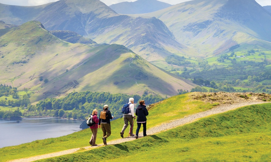 Four walkers on a hillside path above a lake, with green mountains beyond